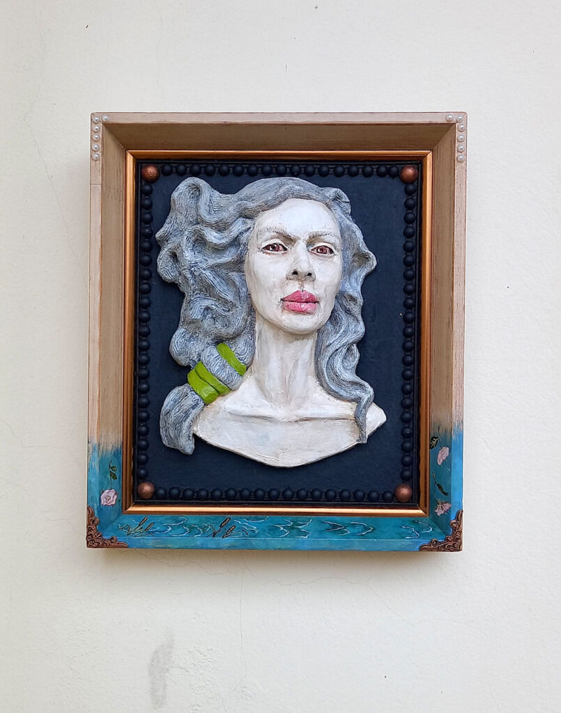 Visão frontal do relevo em cerâmica Vento de Afrodite fixado em moldura com detalhes de pintura na base em fundo branco
