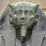 Close-up frontal do rosto do faraó Quéfren com o lenço real Nemés, exibindo uma expressão solene e barba postiça entalhada.