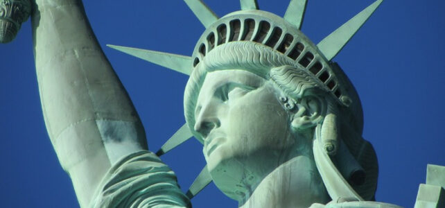 Close-up do rosto da Estátua da Liberdade com as pontas da coroa contra o céu azul.