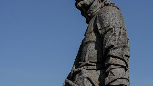 Estátua em pedra-sabão do profeta Joel segurando um pergaminho com inscrições, posicionada no adro do santuário.