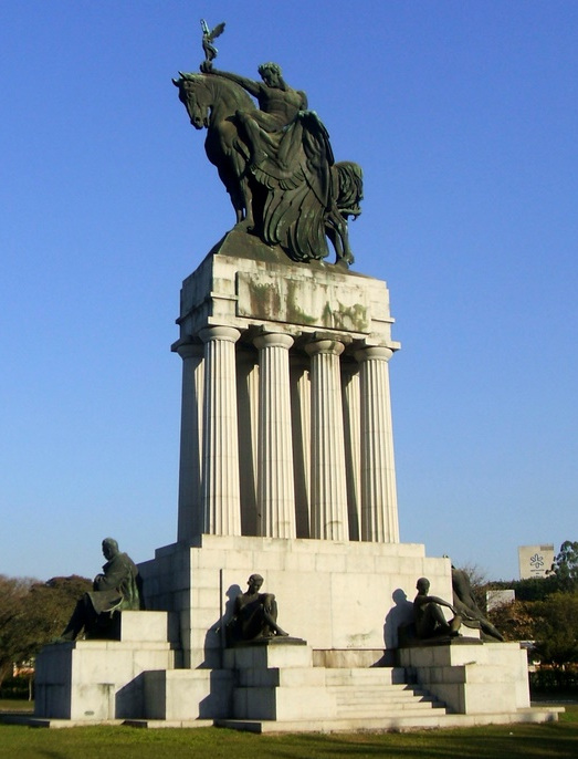 foto de um grande monumento com colunas altas e escultura de bronze de um homem a cavalo na parte superior e nas escadarias alegorias femininas em bronze em um parque sob o céu azul