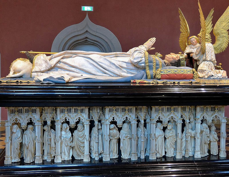 túmulo de um rei muito decorado e colorido com anjos de asas douradas na cabeça do rei deitado e na base muitas esculturas de santos e figuras chorando esculpidas na madeira