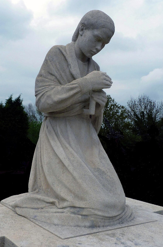estátua delicada feita em mármore branco de uma jovem ajoelhada em um túmulo com as mãos segurando um vela