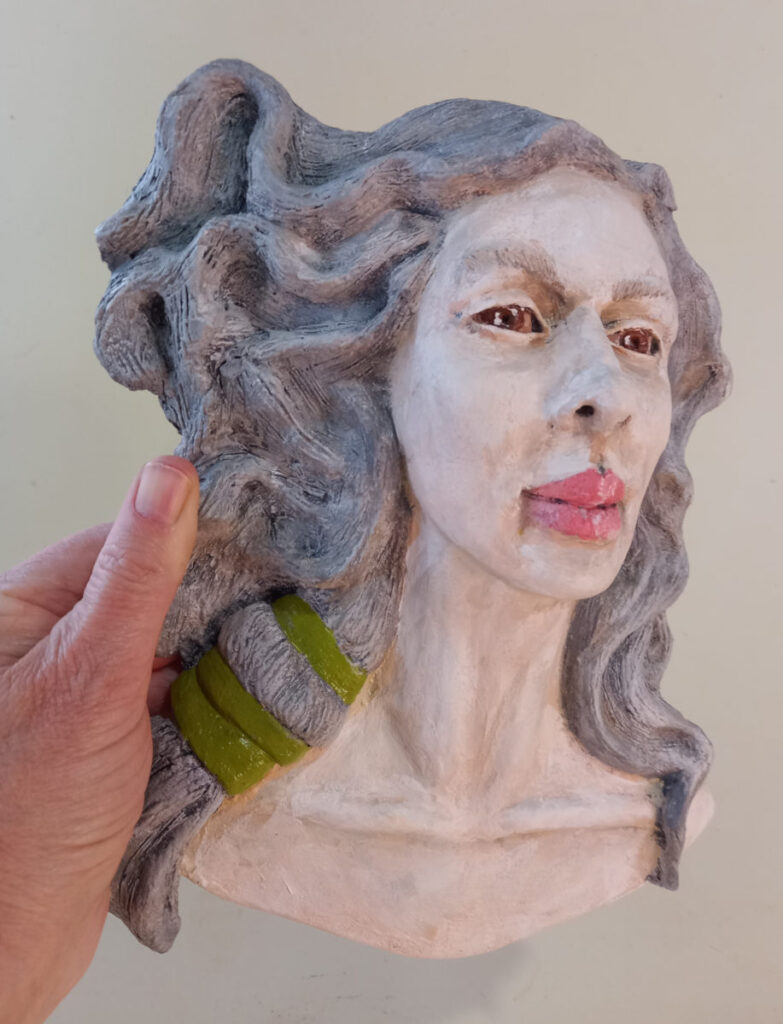 mão segurando uma escultura da cabeça de uma mulher em relevo com cabelos grisalhos, um laço verde nos cabelos e uma expressão tranquila