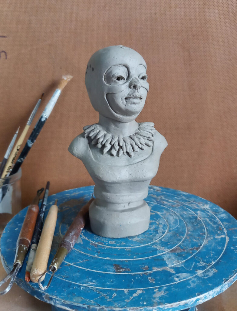 escultura de argila de um busto feminino usando capacete antigo e colar em um ateliê