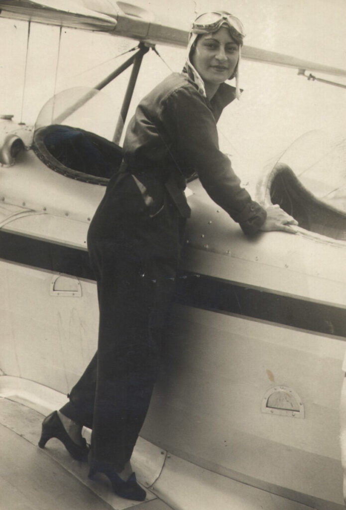 foto em preto e branco de uma aviadora vestida com macacão, capacete e óculos de aviador vintage ao lado de um pequeno avião